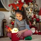 Child wearing reindeer antlers playing with a glowing chick toy in front of a decorated Christmas tree.