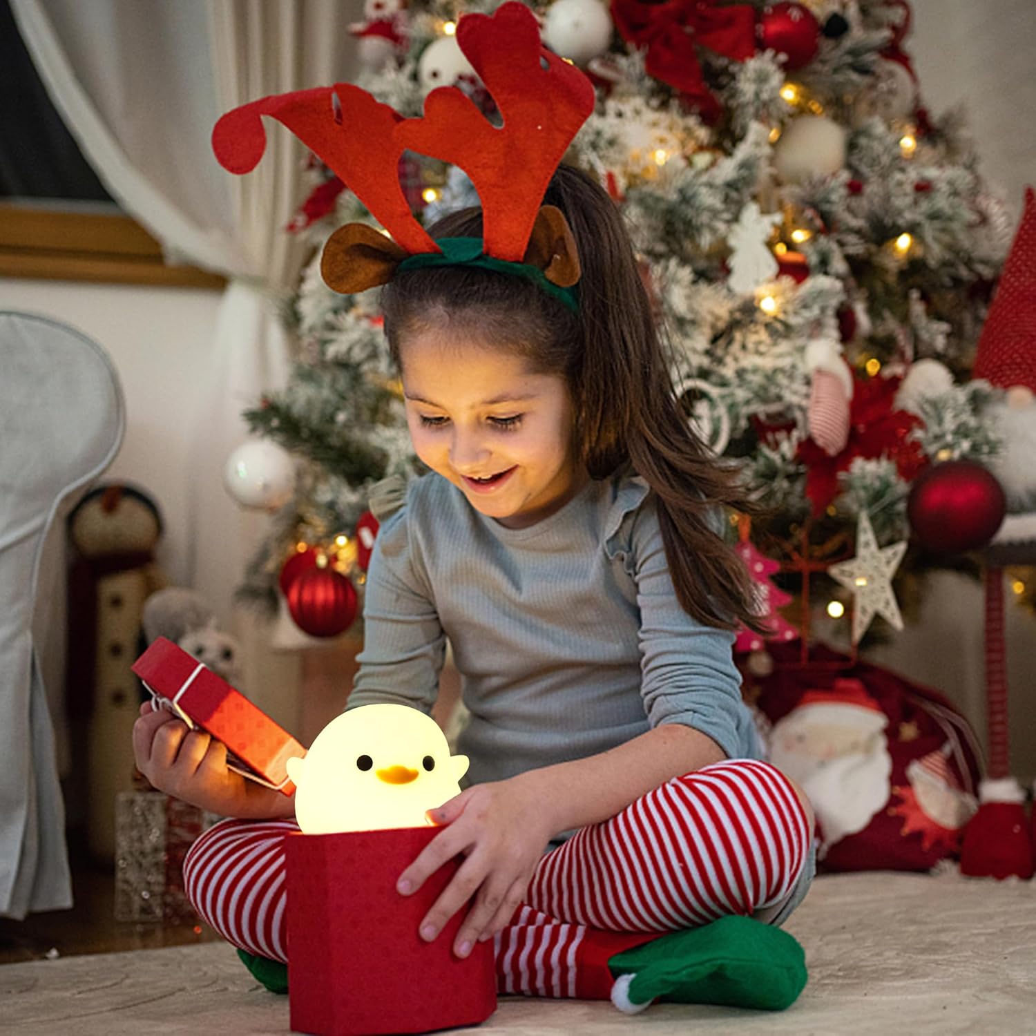 Child wearing reindeer antlers playing with a glowing chick toy in front of a decorated Christmas tree.