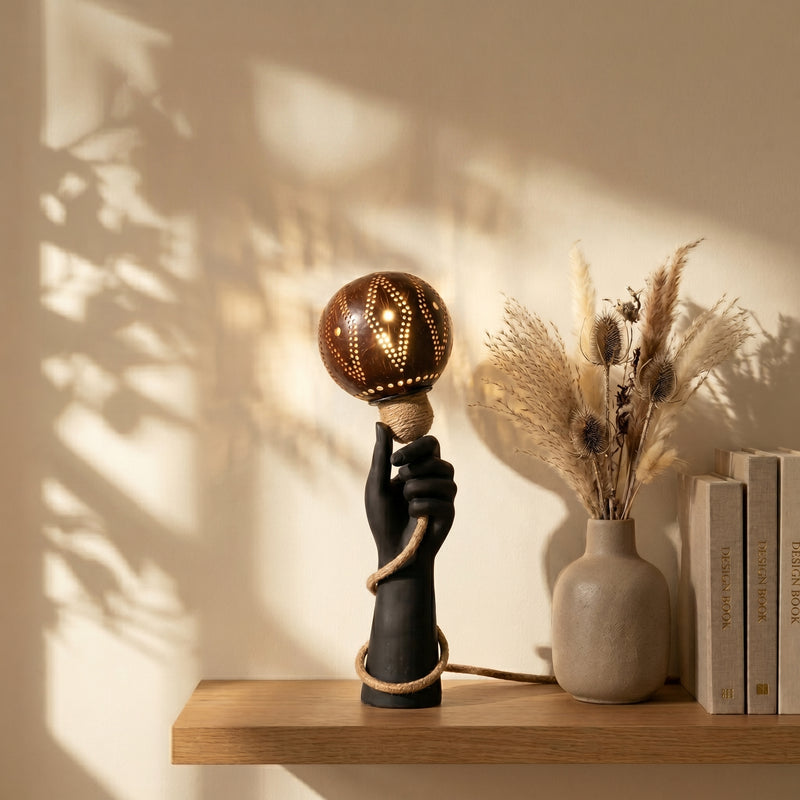 Decorative lamp with a hand holding a bulb on a wooden shelf with books and a vase.